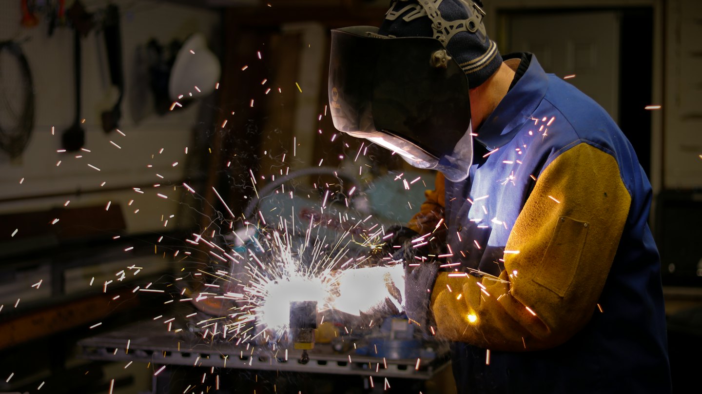 Welder in mask sparks metal on a workbench.